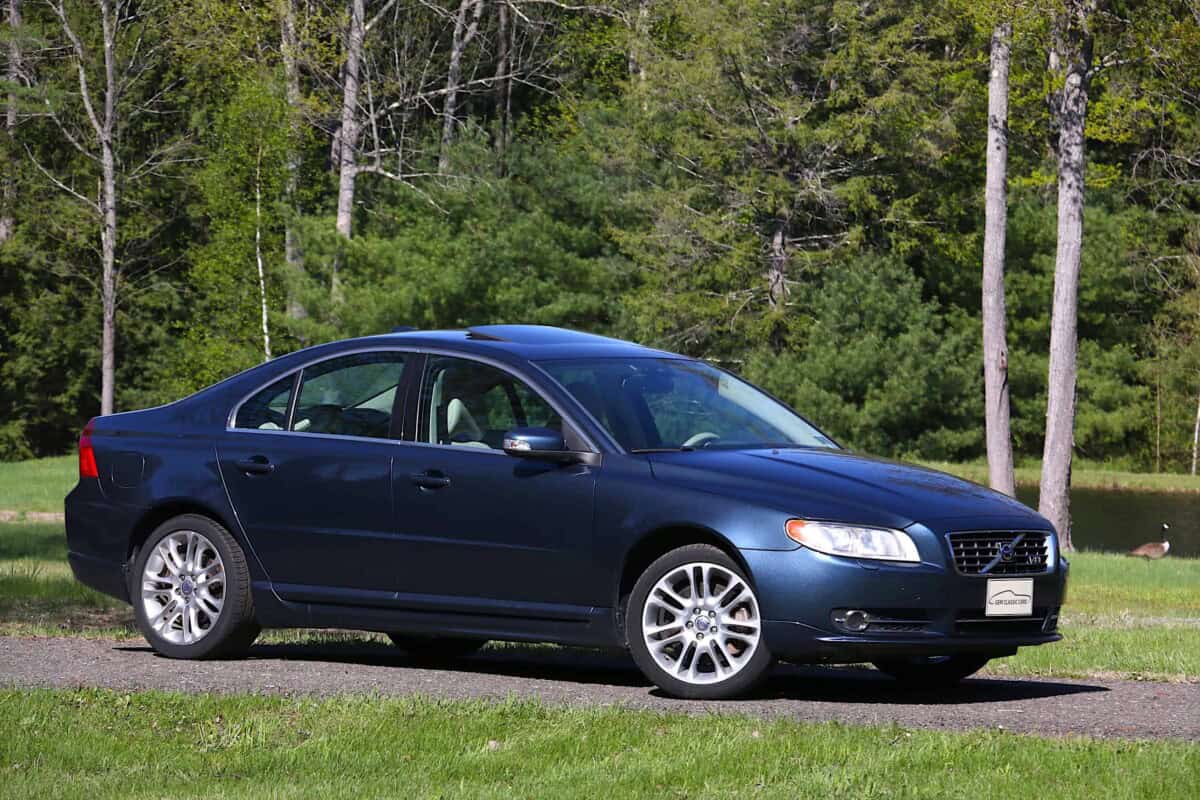 A side view of a blue sedan parked on grass with trees in the background.