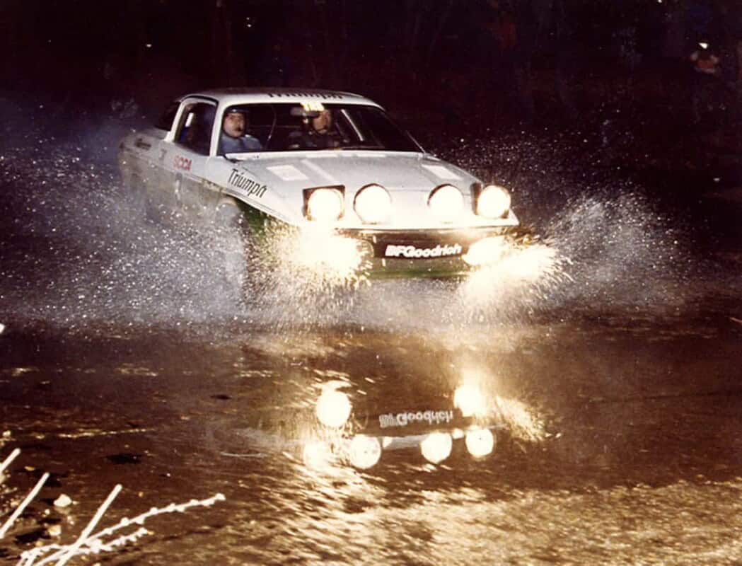 A vintage Triumph rally car driving through water at night, splash and reflectio.