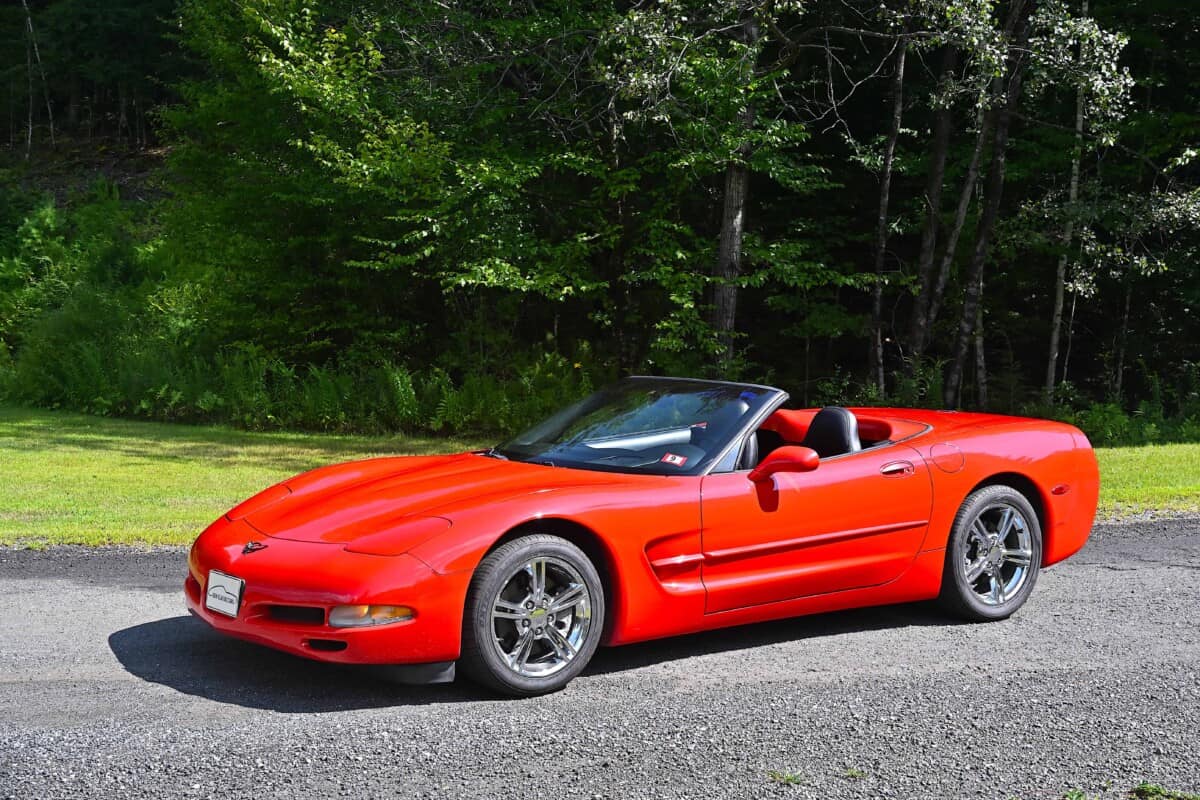 A red Chevrolet Corvette convertible parked on a gravel road with a forest backg.