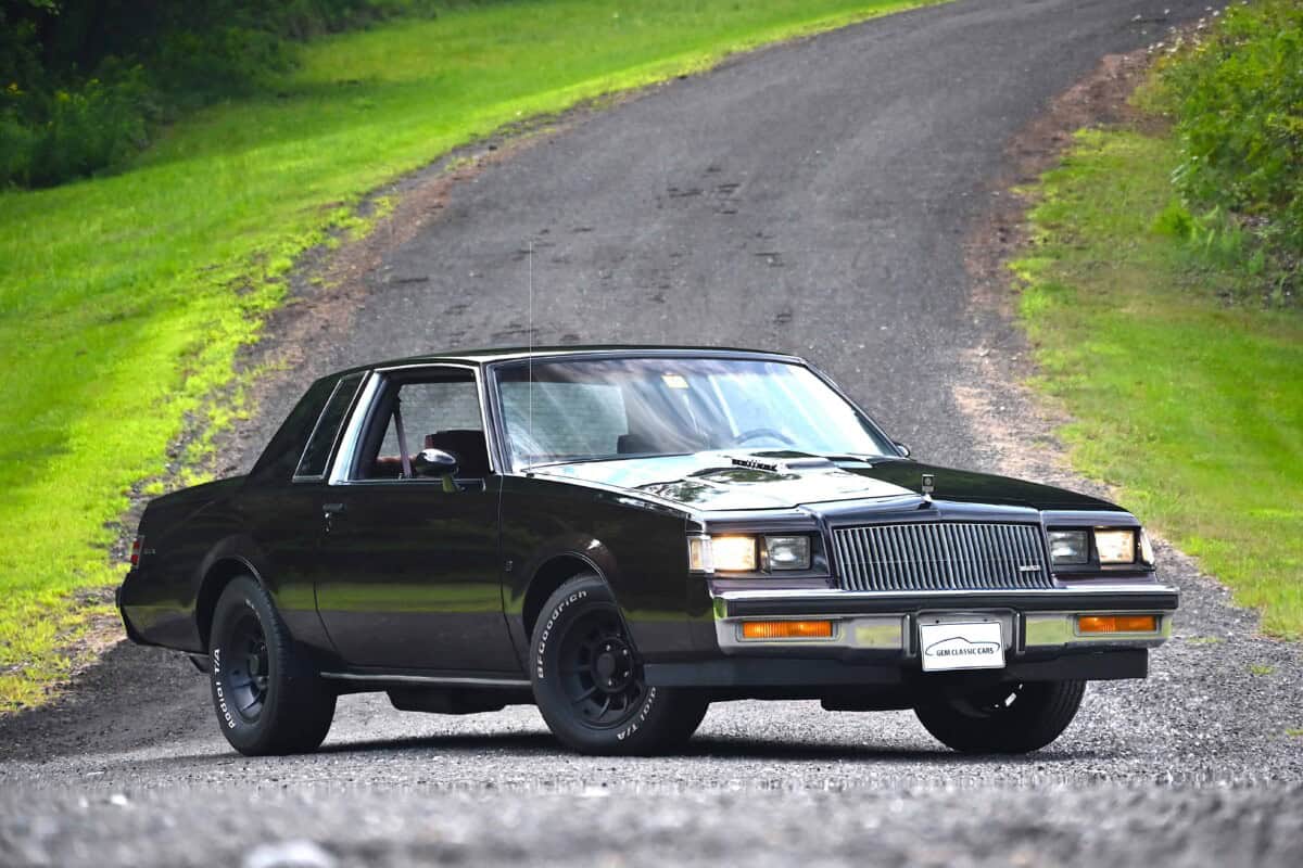 A black vintage luxury sedan parked on a gravel road in a green landscape.