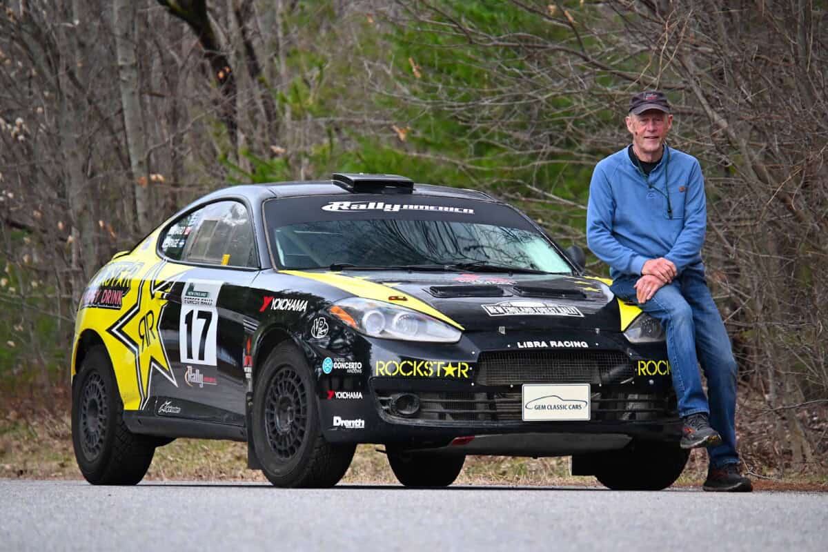 Rally car parked on road with man sitting beside it in outdoor setting.