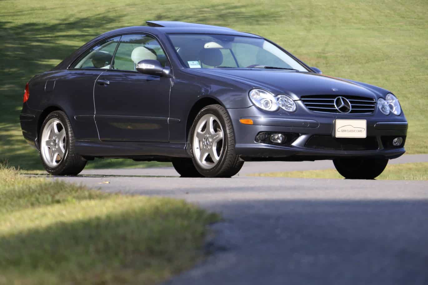 1999 Mercedes CLK500 — Side view of a dark blue classic car coupe on road with grassy background.