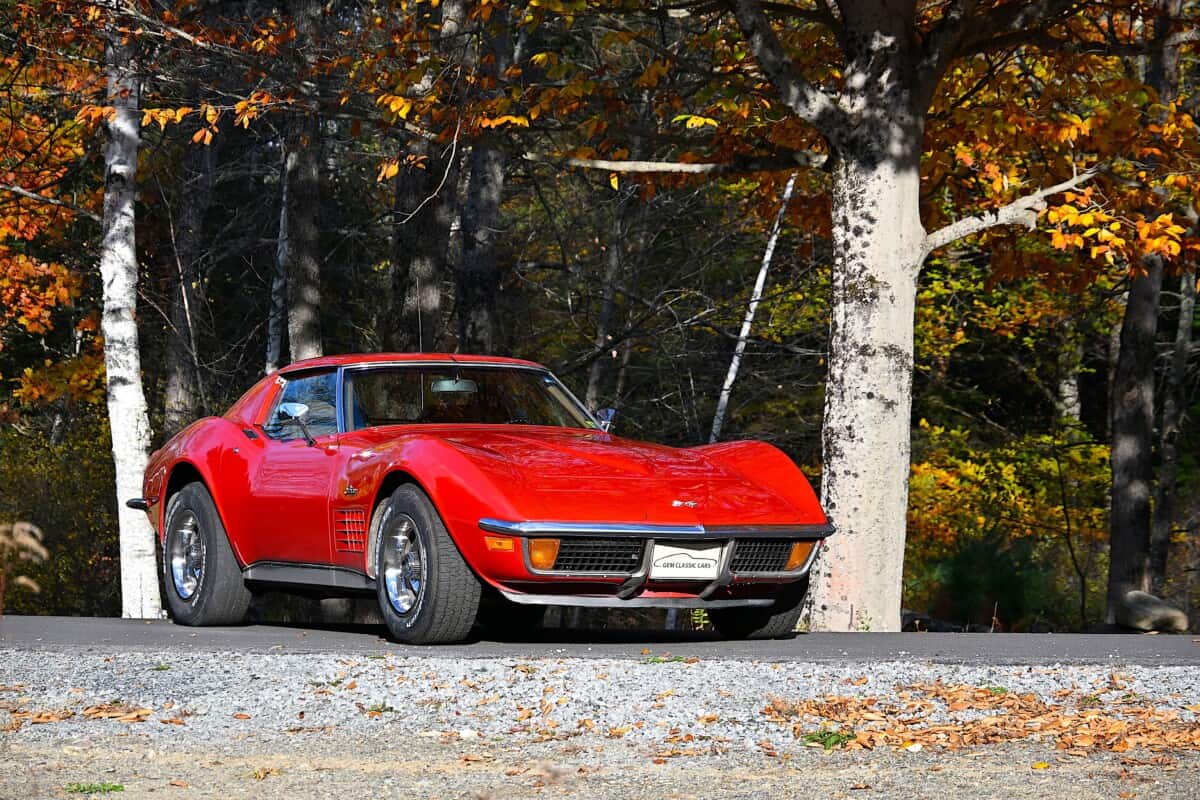 Red classic car parked on a road with autumn trees in background.